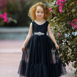 Smiling girl wearing a black sleeveless lace and tulle mermaid-style flower girl dress with a jeweled sash and floral headband, standing near vibrant pink flowers.