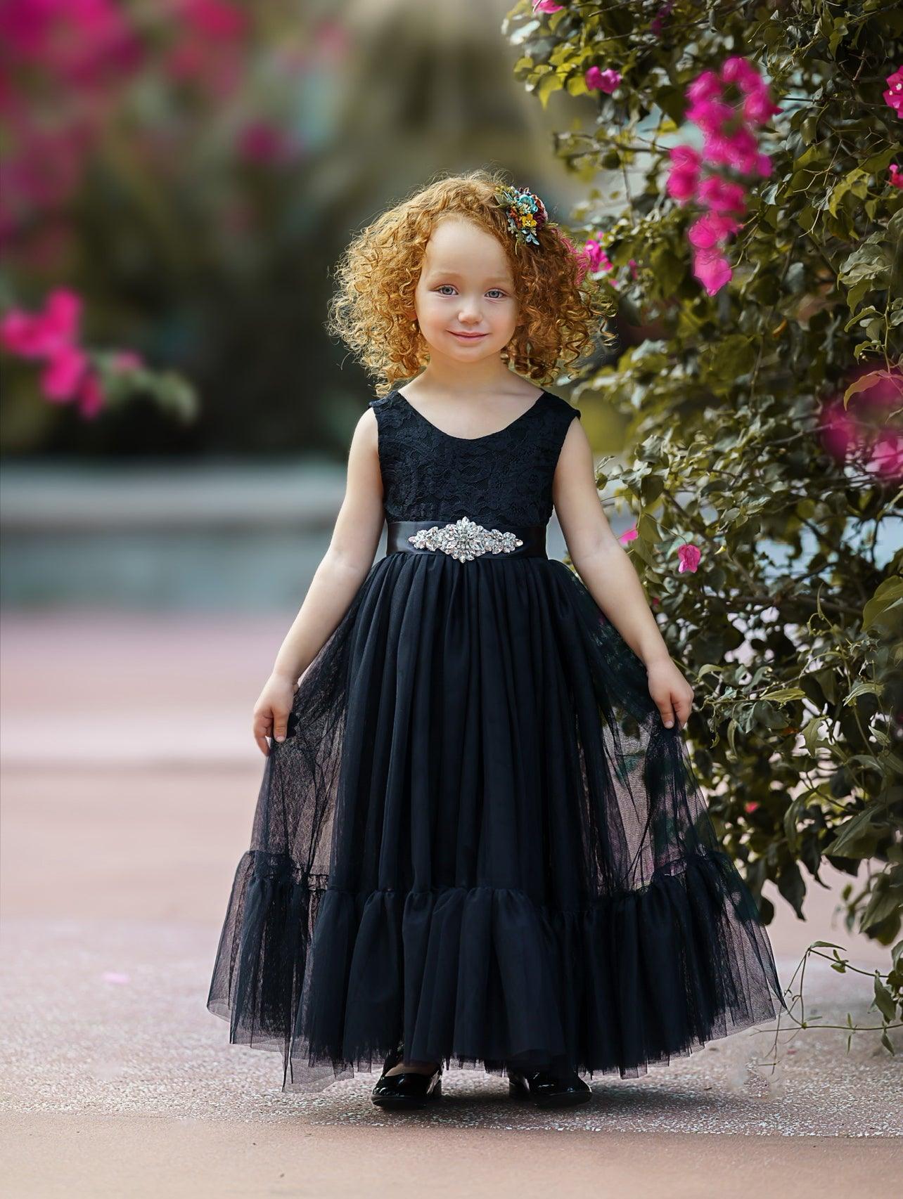 Smiling girl wearing a black sleeveless lace and tulle mermaid-style flower girl dress with a jeweled sash and floral headband, standing near vibrant pink flowers.