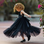Young girl twirling joyfully in a sleeveless black lace and tulle mermaid-style flower girl dress with a satin sash, captured mid-spin outdoors by blooming flowers.