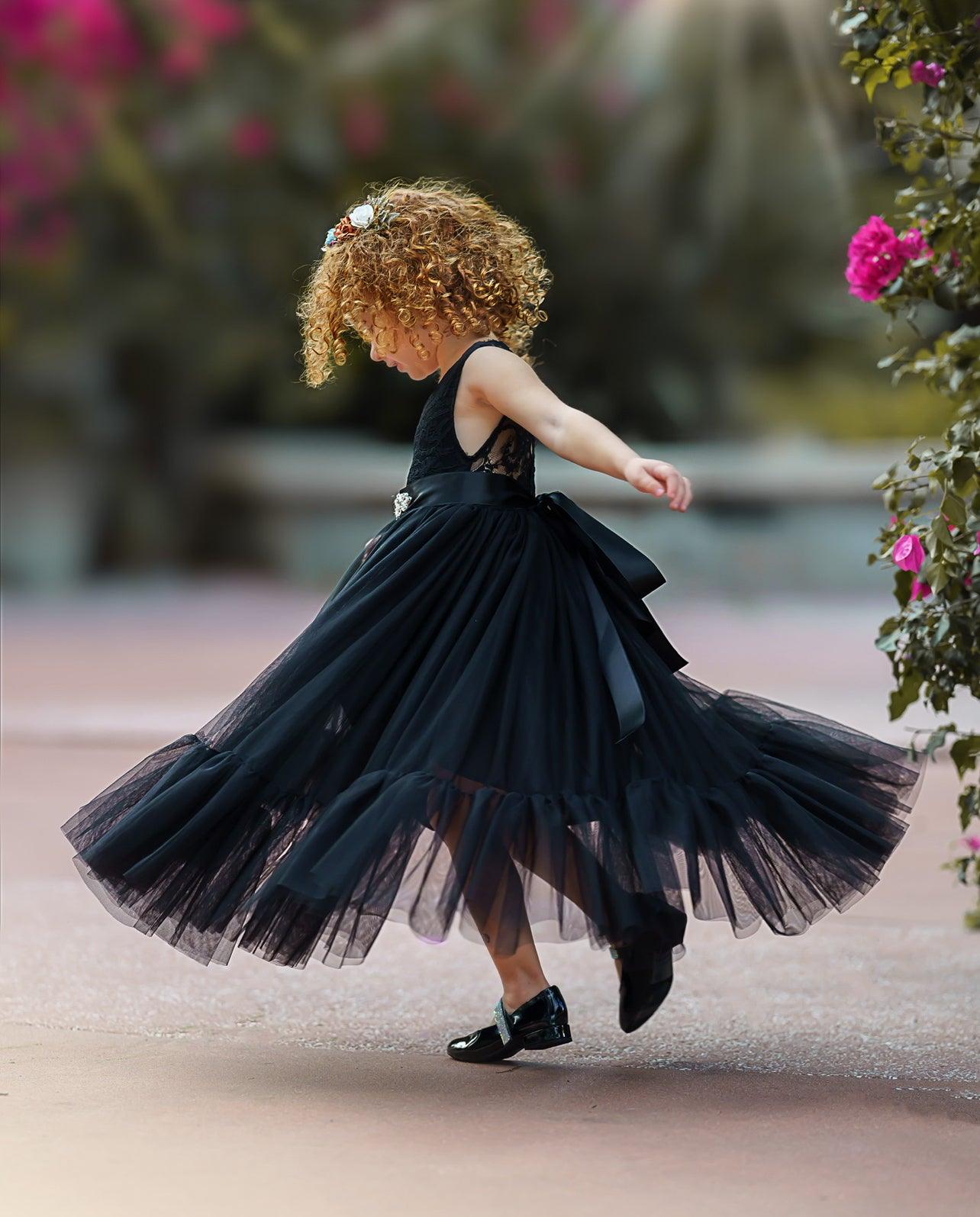 Young girl twirling joyfully in a sleeveless black lace and tulle mermaid-style flower girl dress with a satin sash, captured mid-spin outdoors by blooming flowers.