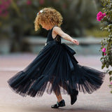 Young girl twirling joyfully in a sleeveless black lace and tulle mermaid-style flower girl dress with a satin sash, captured mid-spin outdoors by blooming flowers.